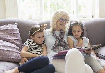 Grandmother and granddaughters reading book on living room sofa - CAIF08908