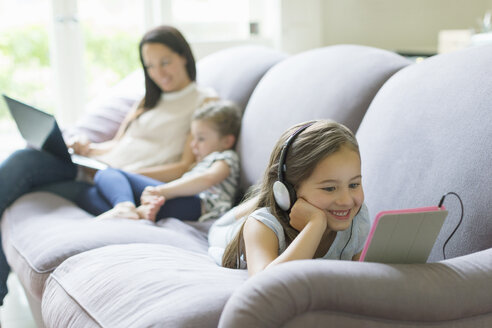 Mädchen mit Kopfhörern und digitalem Tablet auf dem Wohnzimmersofa liegend - CAIF08916