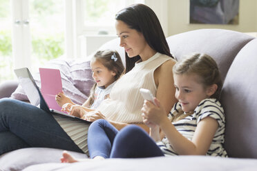 Mother and daughters with laptop, cell phone and digital tablet on sofa - CAIF08922