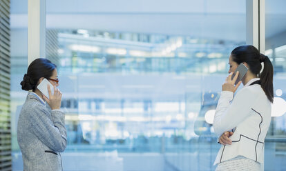 Businesswomen talking on cell phones at office window - CAIF08999