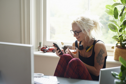 Junge Frau mit Kopfhörern und Tattoo, die im Home Office eine SMS schreibt, lizenzfreies Stockfoto