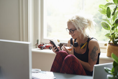 Junge Frau mit Kopfhörern und Tattoo, die im Home Office eine SMS schreibt - CAIF09006