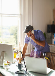 Mann spricht am Telefon und benutzt einen Computer am Schreibtisch im sonnigen Heimbüro - CAIF09013