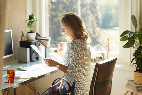 Frau trinkt Kaffee und liest Papierkram am Schreibtisch im sonnigen Heimbüro, lizenzfreies Stockfoto