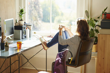 Pensive woman looking through window with feet up on desk in sunny home office - CAIF09028