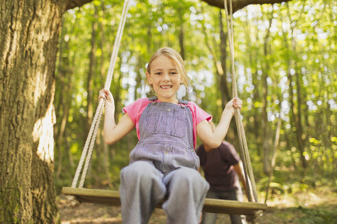 Porträt lächelndes Mädchen schwingt auf Seilschaukel im Wald - CAIF09137