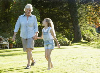 Grandmother and granddaughter holding hands and walking in sunny garden - CAIF09201