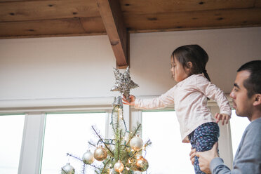 Vater hebt kleines Mädchen hoch, das einen Stern auf den Weihnachtsbaum steckt - CAIF09226