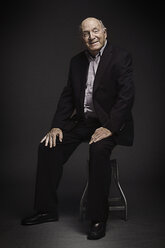 Portrait of senior man sitting on stool against black background - CAVF04087