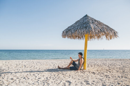 Kuba, Junger Mann sitzt unter einem Sonnenschirm am Playa Ancon - GUSF00543