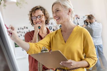 Lächelnde weibliche Designprofis mit Klemmbrett, die im Büro am Flipchart arbeiten - CAIF09238