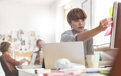 Serious female designer reviewing adhesive notes on computer in office - CAIF09267