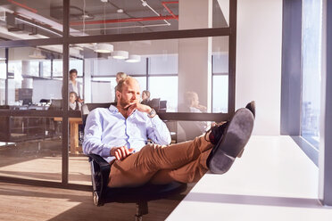 Pensive businessman sitting with feet up looking through sunny window in office - CAIF09295