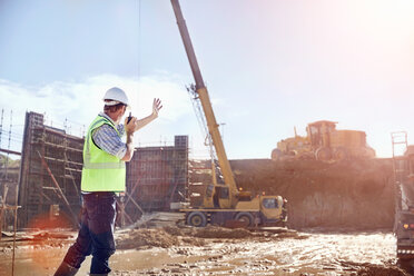 Bauarbeiter, der mit einem Walkie-Talkie einen Kran auf einer sonnigen Baustelle steuert - CAIF09307