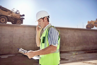 Construction worker foreman talking on cell phone at sunny construction site - CAIF09314