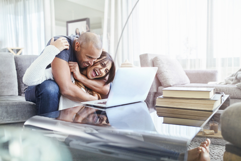 Verspieltes Paar, das sich umarmt und einen Laptop im Wohnzimmer benutzt, lizenzfreies Stockfoto