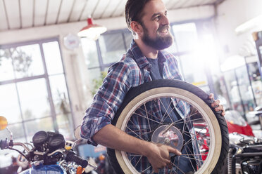 Lächelnder Motorradmechaniker mit Rad in der Werkstatt - CAIF09341