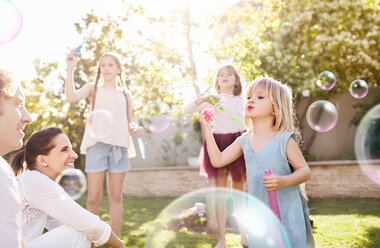 Parents watching daughters blowing bubbles in sunny back yard - CAIF09393