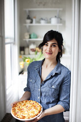 Portrait of woman holding pie while leaning by wall at home - CAVF04479