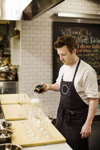 Mann gießt Wein in Gläser ein, während er in einer Großküche steht, lizenzfreies Stockfoto