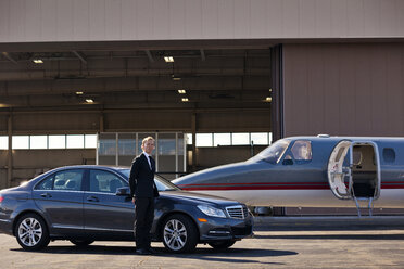 Porträt eines Geschäftsmannes, der neben einem Auto und einem Flugzeug auf einem Flugplatz steht - CAVF04573
