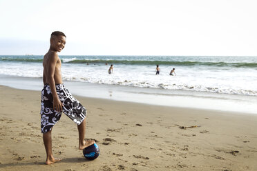Porträt eines Jungen, der mit einem Bein auf einem Fußball am Strand steht - CAVF04837
