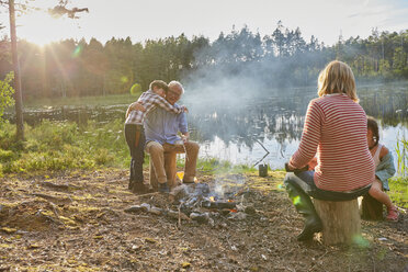 Großeltern und Enkelkinder umarmen sich am Lagerfeuer am sonnigen Seeufer - CAIF09414