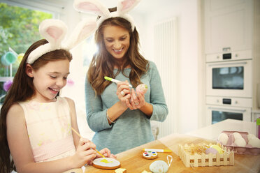 Mutter und Tochter mit kostümierten Hasenohren färben Ostereier und Kekse - CAIF09550