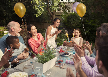 Multiethnische Mehrgenerationen-Familie feiert klatschend Geburtstag mit Feuerwerkskuchen am Terrassentisch - CAIF09562