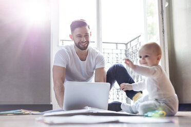 Father working on laptop and watching baby daughter playing on floor - CAIF09656