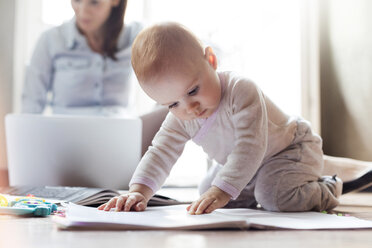 Ein kleines Mädchen spielt auf dem Boden in der Nähe der Mutter, die an einem Laptop arbeitet - CAIF09672