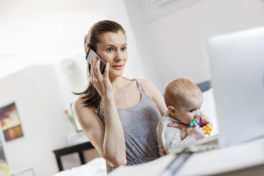 Mutter hält ihre kleine Tochter, arbeitet am Laptop und spricht mit dem Handy - CAIF09678