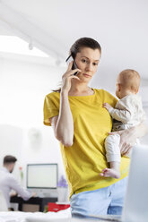 Mother holding baby daughter and talking on cell phone at laptop - CAIF09681