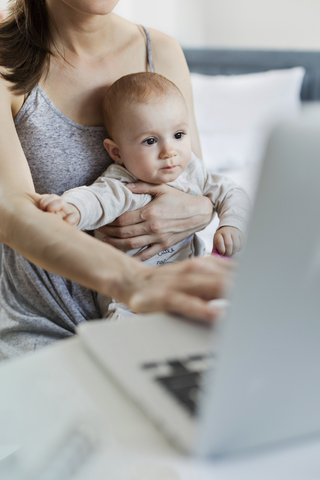 Mutter hält ihre kleine Tochter und arbeitet am Laptop, lizenzfreies Stockfoto