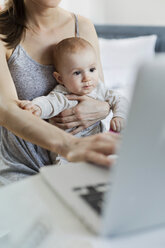 Mother holding baby daughter and working at laptop - CAIF09685