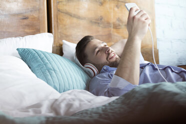Mann hört Musik mit Kopfhörern und mp3-Player auf dem Bett liegend - CAIF09688