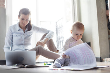 Neugierige Baby-Tochter schaut auf Papierkram neben der Mutter, die am Laptop arbeitet - CAIF09708