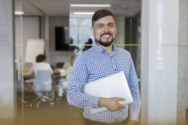 Portrait lächelnder Geschäftsmann mit Laptop im Büro - CAIF09724