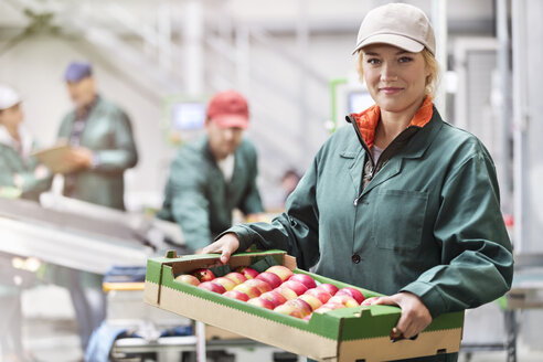 Porträt einer selbstbewussten Arbeiterin, die eine Kiste mit Äpfeln in einem Lebensmittelverarbeitungsbetrieb trägt - CAIF09980