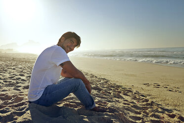Porträt eines lächelnden Mannes auf Sand am Strand sitzend - CAVF04918