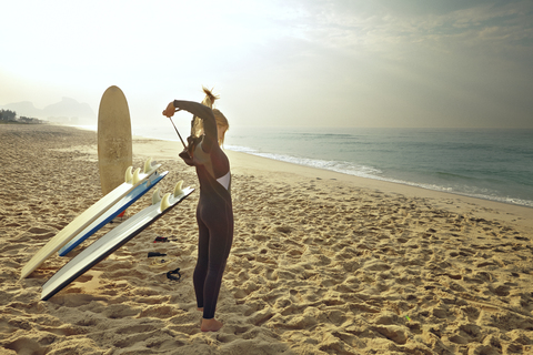 Frau, die ihren Neoprenanzug zuzieht, während sie am Strand bei den Surfbrettern steht, lizenzfreies Stockfoto