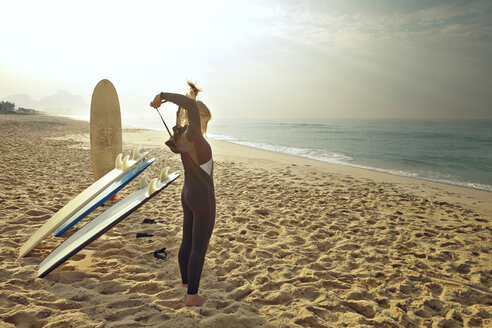 Frau, die ihren Neoprenanzug zuzieht, während sie am Strand bei den Surfbrettern steht - CAVF04920