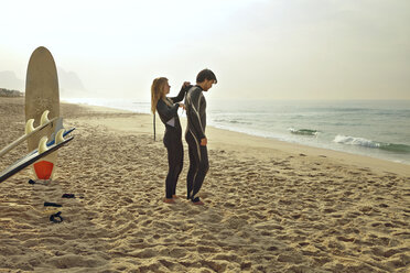 Woman assisting man in wearing wetsuit while standing at beach - CAVF04921