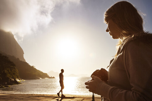 Frau hält Kamera, während Mann am Meer gegen den Himmel steht - CAVF04939