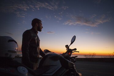 Mann steht bei einem Motorrad und betrachtet die Aussicht bei Sonnenuntergang - CAVF04967