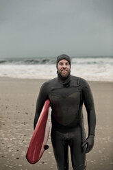 Porträt eines glücklichen Mannes, der ein Surfbrett trägt und am Strand steht - CAVF05089