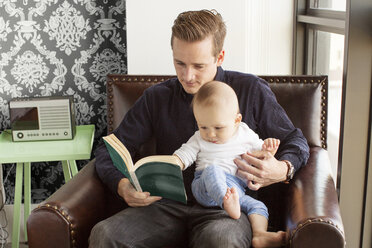 Vater liest mit seinem Sohn ein Buch, während er zu Hause auf einem Sessel sitzt - CAVF05102