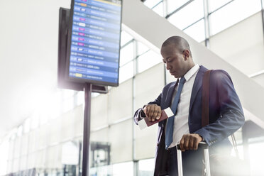 Businessman checking the time on wristwatch below arrival departure board at airport - CAIF10048