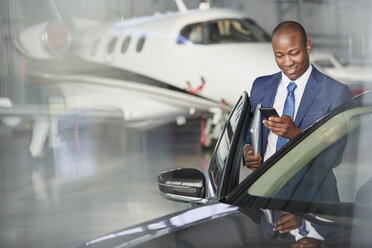 Geschäftsmann mit Mobiltelefon steigt in der Nähe des Firmenjets im Hangar ins Auto - CAIF10053