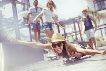 Porträt einer verspielten jungen Frau, die auf der sonnigen Strandpromenade Skateboard fährt - CAIF10145